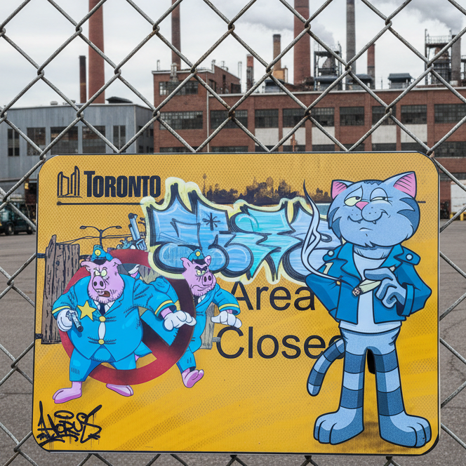 Cartoon-style sign with characters and text on a chain-link fence with industrial buildings in the background