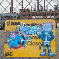 Cartoon-style sign with characters and text on a chain-link fence with industrial buildings in the background